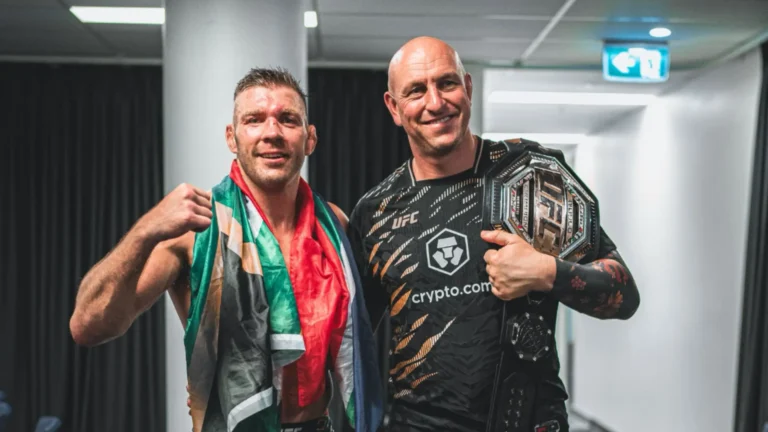 Dricus du Plessis draped in the South African flag beside CoreCraft coach Niel holding the UFC championship belt backstage